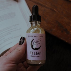 Puede incluir: Una botella de vidrio transparente de "Healer Potion Oil" con un cuentagotas negro, sostenida en una mano. La etiqueta es de color lila claro con un diseño de luna creciente, cristal y planta. La botella contiene 60 ml de aceite y es de Midnight Raven Studios.