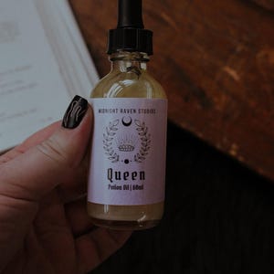 Może przedstawiać: Mała szklana butelka Queen Potion Oil z czarną pipetą. Etykieta jest jasnofioletowa z napisem "Midnight Raven Studios" i "Queen". Butelka zawiera 60 ml olejku.