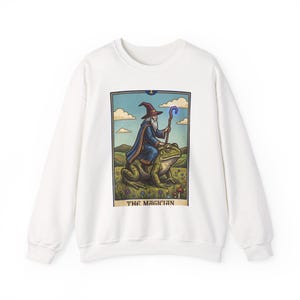 May include: White sweatshirt featuring a tarot card design. The graphic depicts a wizard riding a frog, holding a staff, with the words "THE MAGICIAN" at the bottom. The wizard wears a blue robe and hat.