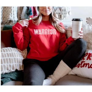 Puede incluir: Una sudadera roja con la palabra "WARRIOR" impresa en blanco en la parte delantera. La sudadera la lleva puesta una persona sentada en un sof&aacute;.