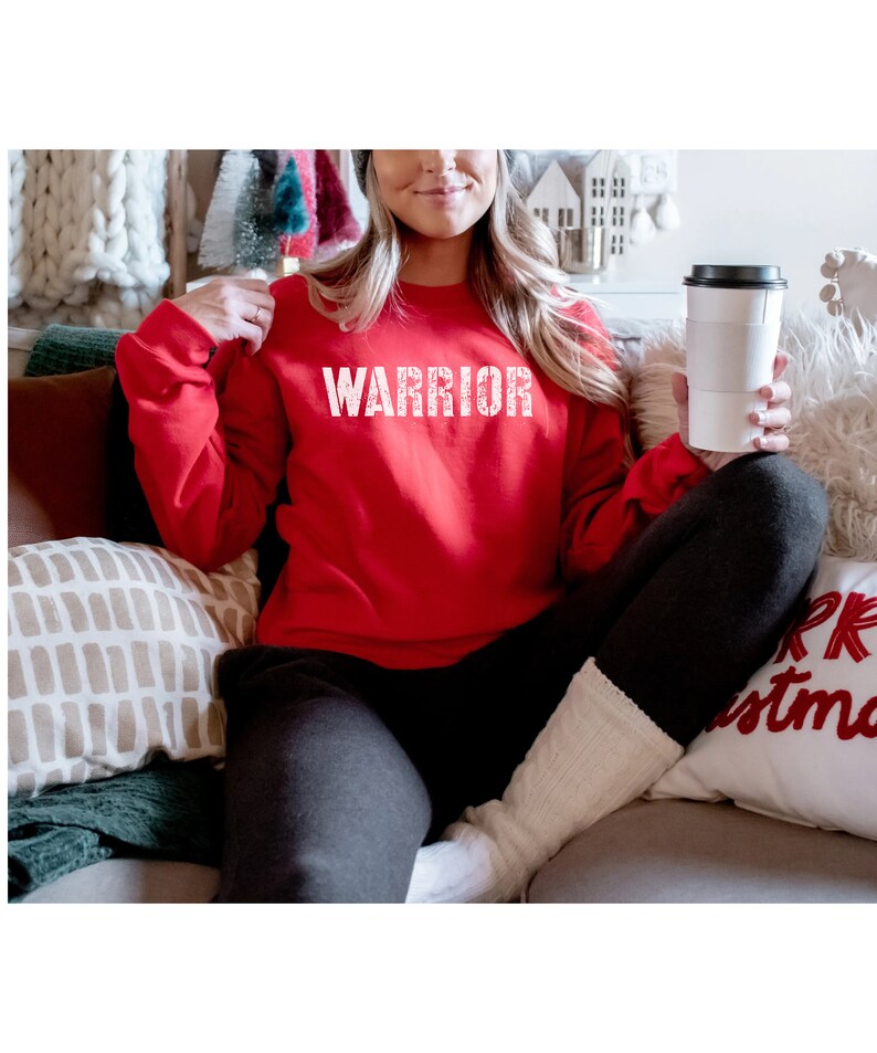 Puede incluir: Una sudadera roja con la palabra "WARRIOR" impresa en blanco en la parte delantera. La sudadera la lleva puesta una persona sentada en un sof&aacute;.