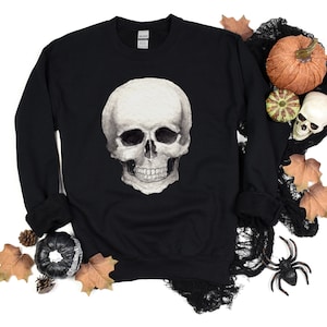 May include: Black sweatshirt with a white skull graphic print. The skull has a detailed design with a white background and black eyes and teeth. The sweatshirt is surrounded by fall-themed decorations, including pumpkins, leaves, and spiderwebs.
