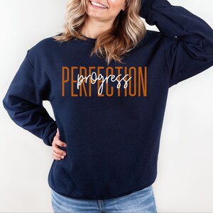 May include: Navy blue sweatshirt with the words "PERFECTION" in orange block letters and "progress" in white cursive script. The sweatshirt is a crew neck style.