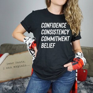 May include: Black t-shirt with the words "CONFIDENCE", "CONSISTENCY", "COMMITMENT", and "BELIEF" printed in white.