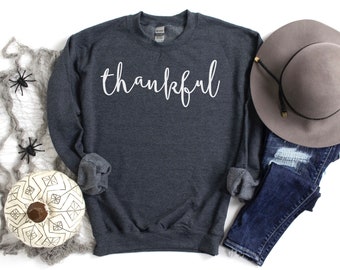 May include: A dark grey sweatshirt with the word "thankful" written in white on the front. The sweatshirt is laid flat on a white surface with a hat, jeans, and a decorative pumpkin.