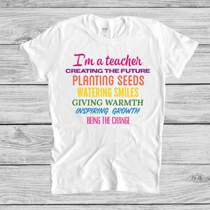 May include: White t-shirt with the text "I'm a teacher" in pink, followed by a list of phrases in rainbow colors: "Creating the future", "Planting seeds", "Watering smiles", "Giving warmth", "Inspiring growth", and "Being the change".