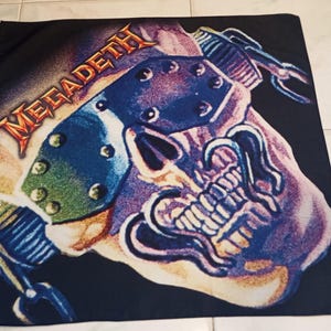 May include: Black square textile featuring a skull graphic with a guitar headstock and the word "MEGADETH" in orange. The design includes blue, purple, and white accents, with a chain detail. The textile is likely a band merchandise item.