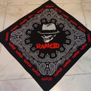 May include: Black bandana with a white paisley design and the word "RANCID" in red. The center features a skull wearing a fedora. The word "RANCID" is also printed around the border of the bandana.