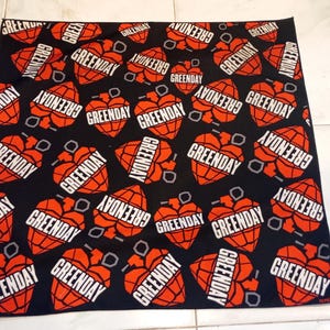 May include: Black fabric with a repeating pattern of red heart-shaped grenades with the word "GREENDAY" in white. The design includes small gray circles. The fabric is likely a bandanna or similar accessory.