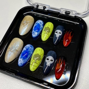 May include: A set of ten artificial nails in a black case. The nails feature various designs, including a shimmering gold, blue water effect, a green design, a skull, and a flame effect. The nails are oval-shaped and have a glossy finish.
