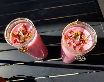 May include: Two pink glass jar candles with a pentagram charm. The candles are topped with dried flowers and herbs.