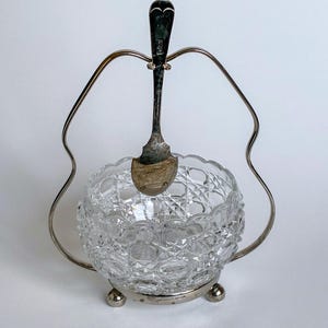 May include: A clear, cut-glass sugar bowl with a silver-colored metal stand and spoon. The bowl has a diamond pattern and sits on three small feet. The spoon rests inside the bowl, held by the stand.