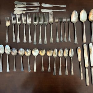 May include: An assortment of vintage silverware, including forks, spoons, knives, and a cake server, arranged on a dark brown surface. The silverware is silver-toned and shows signs of age.