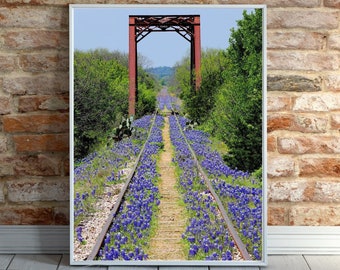 May include: A framed wall art print featuring a perspective view of railroad tracks stretching into the distance, flanked by vibrant bluebonnet flowers. A red metal bridge structure spans the tracks, surrounded by green trees under a clear blue sky.