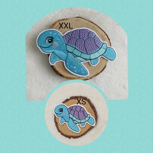 Könnte beinhalten: Zwei gestickte Schildkrötenaufnäher, einer mit der Aufschrift "XXL" und der andere mit "XS". Die Schildkröten haben blaue Körper, lila Panzer und weiße Umrisse. Sie sind auf Holzscheiben vor hellem Hintergrund platziert.
