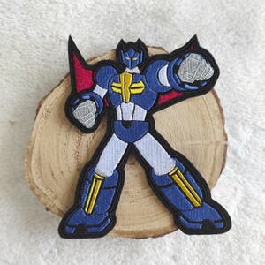 Könnte beinhalten: Eine blaue und gelbe Roboterfigur mit roten Flügeln, gestickt auf einem schwarzen Stoffpatch.
