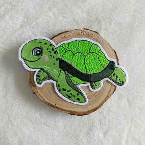 Könnte beinhalten: Grüner gestickter Meeresschildkröten-Patch mit schwarzen Augen und schwarzem Panzerumriss. Der Patch befindet sich auf einem hölzernen Hintergrund.