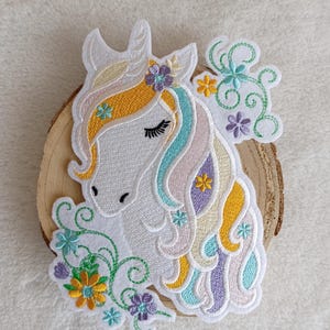 Könnte beinhalten: Gestickter Einhorn-Aufnäher mit weißem Gesicht, geschlossenen Augen und bunter Mähne in Gelb, Blau und Lila. Der Aufnäher hat florale Akzente in verschiedenen Farben und ist auf einer Holzscheibe angebracht.