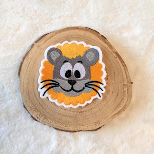 Könnte beinhalten: Nahaufnahme eines gestickten Löwenkopf-Patches mit grauem Gesicht, weißen Augen und gelber Mähne. Der Patch befindet sich auf einem hölzernen Hintergrund.