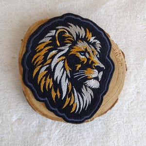 Könnte beinhalten: Gestickter Aufnäher mit einem Löwenkopf im Profil. Die Löwenmähne ist mit goldenen und weißen Fäden detailliert, vor einem schwarzen Hintergrund mit einem marineblauen Rand. Der Löwe hat blaue Augen. Der Aufnäher liegt auf einer Holzscheibe.