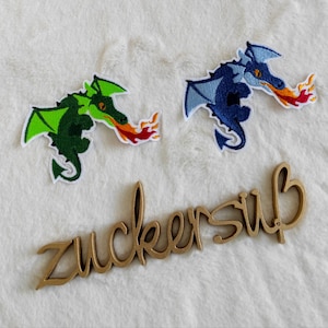 Könnte beinhalten: Zwei gestickte Bügelbilder von Drachen, die Feuer spucken. Der Drache links ist grün und der Drache rechts ist blau. Das Wort "zuckersüß" ist in Gold auf einer Holzoberfläche geschrieben.