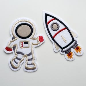 Könnte beinhalten: Zwei gestickte Aufnäher: ein Astronaut in einem weißen Raumanzug mit roten Akzenten und eine weiße Rakete mit roten und schwarzen Details. Der Astronautenaufnäher hat ein beiges Visier und die Rakete hat orangefarbene Flammen.