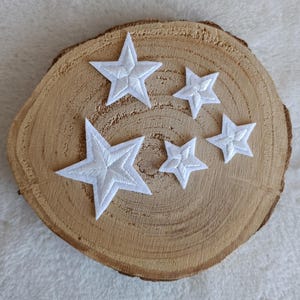 Könnte beinhalten: Fünf weiße, gestickte Sternaufnäher in verschiedenen Größen sind auf einer Holzscheibe angeordnet. Die Sterne haben ein detailliertes, strukturiertes Aussehen mit sichtbaren Nähten. Der Hintergrund ist ein weicher, hellfarbiger Stoff.