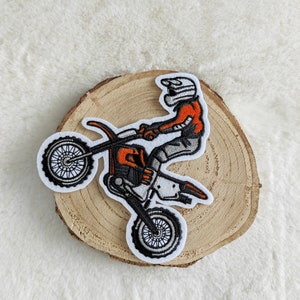 Könnte beinhalten: Ein gestickter Aufnäher mit einem Motocross-Fahrer in orangefarbener und grauer Kleidung, der ein weißes und schwarzes Dirtbike fährt. Der Aufnäher befindet sich auf einem hölzernen Hintergrund.