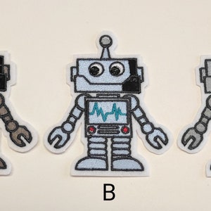 Könnte beinhalten: Drei gestickte Roboter-Patches in verschiedenen Farbvarianten: Braun, Hellblau und Grau. Jedes Patch zeigt einen Roboter mit einem Bildschirm, der einen Herzfrequenzmonitor, eine blaue Antenne und große Augen anzeigt.