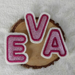 Könnte beinhalten: Drei rosa Filzbuchstaben, die den Namen "EVA" mit Glitzerakzenten buchstabieren. Die Buchstaben befinden sich auf einer Holzscheibe.