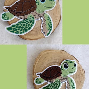 Könnte beinhalten: Zwei aufgestickte Meeresschildkröten-Patches. Die Schildkröten haben braune Panzer, grüne Köpfe und Flossen sowie große, freundliche Augen. Die Patches befinden sich auf einem Holzscheiben-Hintergrund mit einem hellgrünen Hintergrund.