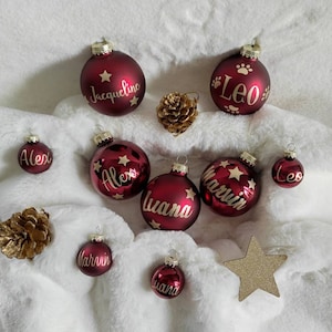 Könnte beinhalten: Burgunderfarbene Weihnachtskugeln mit Namen wie "Jacqueline", "Alex", "Luana", "Marvin" und "Leo" in goldener Schrift. Einige Kugeln haben Sterndesigns. Sie sind auf einer weißen, flauschigen Oberfläche angeordnet, mit goldenen Tannenzapfen und einem Stern.