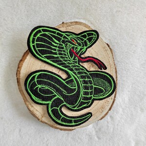 Könnte beinhalten: Grün-schwarzer gestickter Bügel-Patch mit einer Kobra, die ihre Haube erhoben und die Zunge ausgestreckt hat. Der Patch befindet sich auf einer Holzoberfläche.