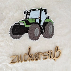 Könnte beinhalten: Ein gestickter Aufnäher eines grünen und schwarzen Traktors mit großen grauen Reifen. Der Traktor hat eine hellblaue Windschutzscheibe und einen weißen Rand. Darunter das Wort "zuckersüß" aus Holz.
