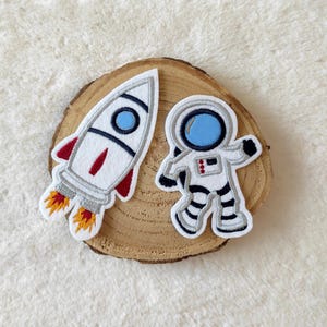 Könnte beinhalten: Zwei gestickte Aufnäher: eine weiße Rakete mit roten und orangen Flammen und ein weißer Astronaut mit schwarzen und grauen Details. Beide Aufnäher haben blaue Akzente und sind auf einer Holzscheibe platziert.
