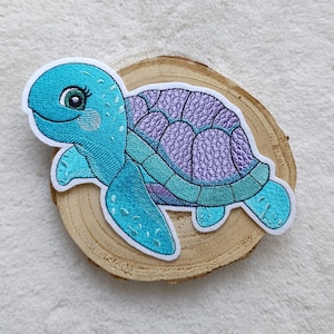 Könnte beinhalten: Gestickter Aufnäher einer lächelnden Cartoon-Schildkröte. Die Schildkröte hat einen hellblauen Kopf und Flossen, einen lila Panzer und einen weißen Rand. Der Aufnäher befindet sich auf einer Holzscheibe.