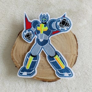 Könnte beinhalten: Ein gestickter Bügelaufkleber mit einem blauen, grauen und gelben Roboter mit einem roten Umhang. Der Roboter befindet sich in Kampfstellung mit ausgestreckten Armen.