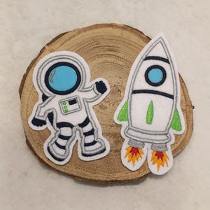 Könnte beinhalten: Zwei Filzpatches, einer mit einem Astronauten in einem weißen Raumanzug mit blauem Helm und der andere mit einer weißen Rakete mit grünen Flammen, auf einem Holzgrund.