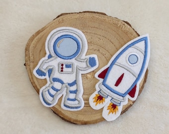 May include: Two embroidered iron-on patches, one of an astronaut in a white spacesuit with a blue helmet and the other of a blue and white rocket with red flames.