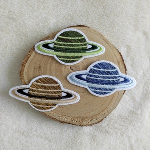 Könnte beinhalten: Drei gestickte Bügelbilder von Saturn, jedes mit einem anderen Farbring: grün, blau und braun.