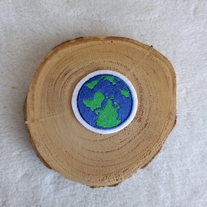 Könnte beinhalten: Gestickter Aufnäher mit einem stilisierten Erdendesign. Der Aufnäher hat einen blauen Rand und einen weißen Hintergrund. Die Kontinente sind grün und die Ozeane blau. Der Aufnäher liegt auf einer Holzscheibe.
