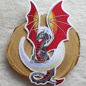 Könnte beinhalten: Gestickter Drachen-Aufnäher mit einem rot-gelben geflügelten Drachen mit grauem Körper und einem weißen Halbmond. Der Aufnäher befindet sich auf einer Holzscheibe, ideal für Kleidung oder Accessoires.