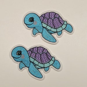 Könnte beinhalten: Zwei gestickte Bügelbilder von blauen und violetten Meeresschildkröten. Die Schildkröten haben ein lächelndes Gesicht und schwimmen nach rechts.