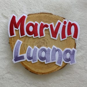 Könnte beinhalten: Gestickte Aufnäher mit den Namen "Marvin" und "Luana" in Rot bzw. Lila, jeweils mit weißen Umrandungen. Die Aufnäher sind auf einer natürlichen Holzscheibe platziert, was eine rustikale Ästhetik erzeugt. Der Text ist in einer verspielten, stilisierten Schriftart.