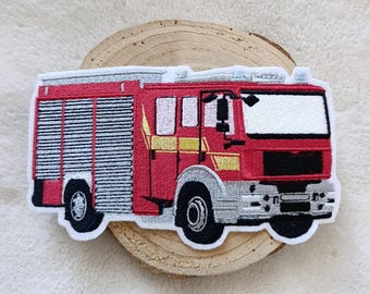 Puede incluir: Parche bordado de un camión de bomberos rojo con detalles plateados y negros. El parche presenta un diseño detallado del vehículo, incluyendo ventanas, ruedas y una escalera. El parche está sobre una rodaja de madera.