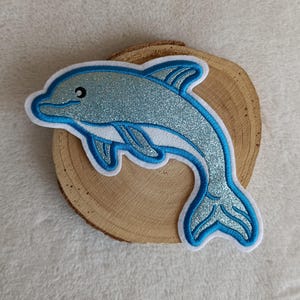 Könnte beinhalten: Ein blauer Glitzer-Delfin-Aufnäher mit weißem Rand und hellblauer Umrandung. Der Delfin hat ein lächelndes Gesicht und ist vor einem Holzgrund platziert. Ideal zum Aufnähen auf Kleidung oder Accessoires.