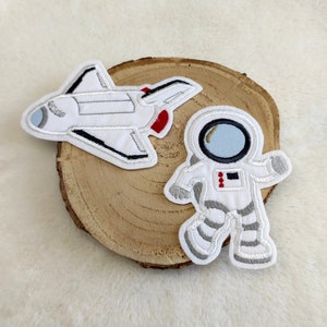 Könnte beinhalten: Zwei gestickte Bügelbilder, eines mit einem weißen Raumschiff und eines mit einem weißen Astronauten mit blauem Visier.