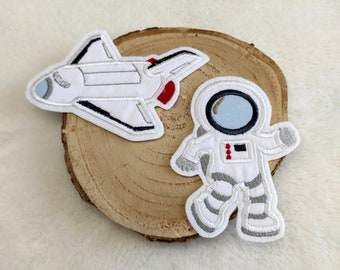 May include: Two embroidered iron-on patches, one of a white spaceship and one of a white astronaut with a blue visor.