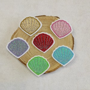 Könnte beinhalten: Sechs glitzernde Patches in Muschelform in verschiedenen Farben, darunter Gold, Rosa, Rot, Blau, Lila und Grün, auf einer Holzoberfläche angeordnet.