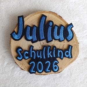 Könnte beinhalten: Eine kreisförmige Holzscheibe mit einem gestickten Aufnäher, der den Text Julius Schulkind 2026 zeigt. Die Buchstaben sind in hellblauem Garn mit einem dicken schwarzen Rand gestickt, was einen starken Kontrast zur natürlichen Holzmaserung bildet.